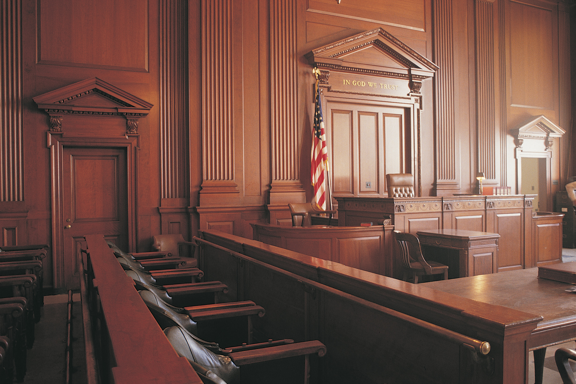 An empty courtroom with wooden benches, a judge’s bench, and an American flag.